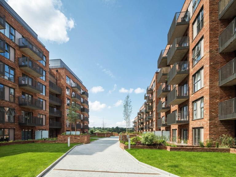 Photo of Springfield Park in Maidstone. Pathway with grass either side between two brown brick apartment buildings; balconies overlooking the pathway. Blue sky with white clouds.